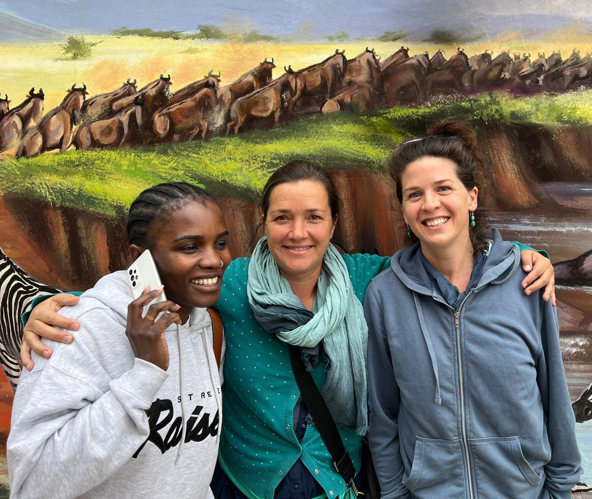 Hanna Boesch umarmt Lovina Kainrath und Nora Breitenecker vor einer, mit einer afrikanischen Szene bemalten, Wand